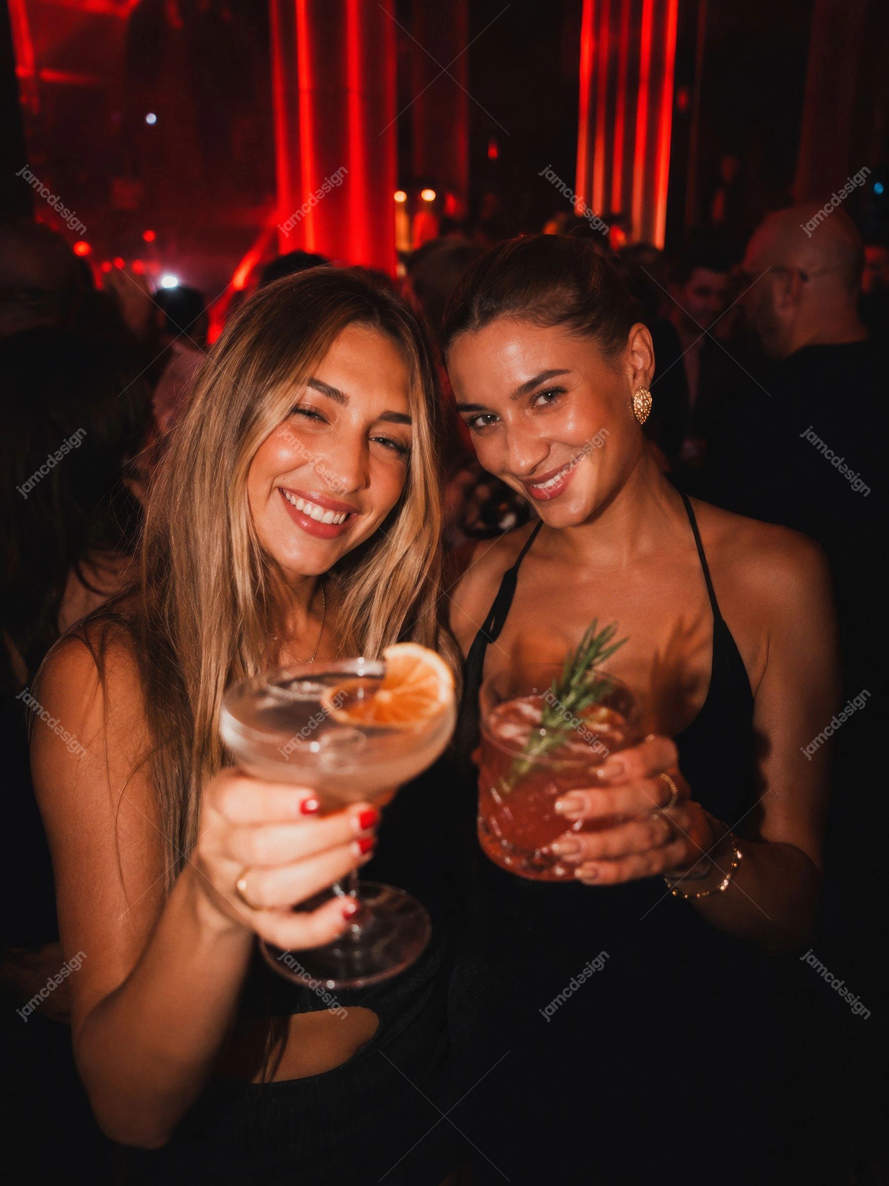 Foto De Amigas Com Coquetéis Em Festa De Boate Vermelha. Foto JPG de duas mulheres segurando taças de coquetel em uma boate lotada