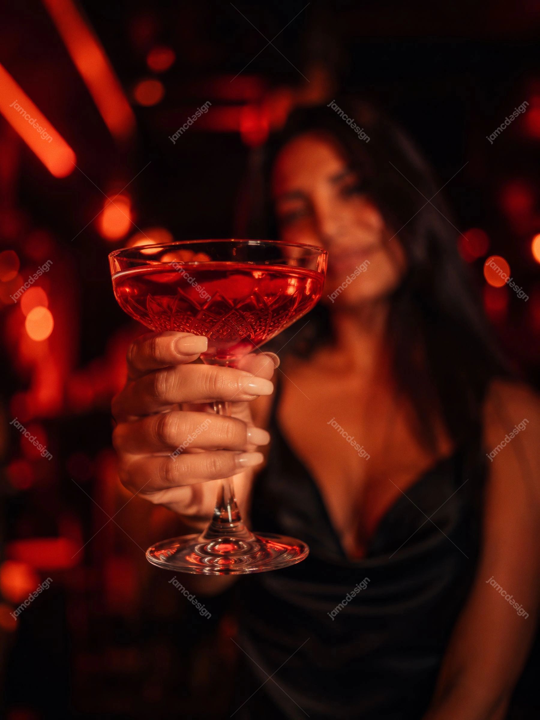 Coquetel Vermelho Em Taça Coupe Em Destaque Com Mulher Ao Fundo. Foto em JPG de um coquetel vermelho em taça coupe, segurado em primeiro plano
