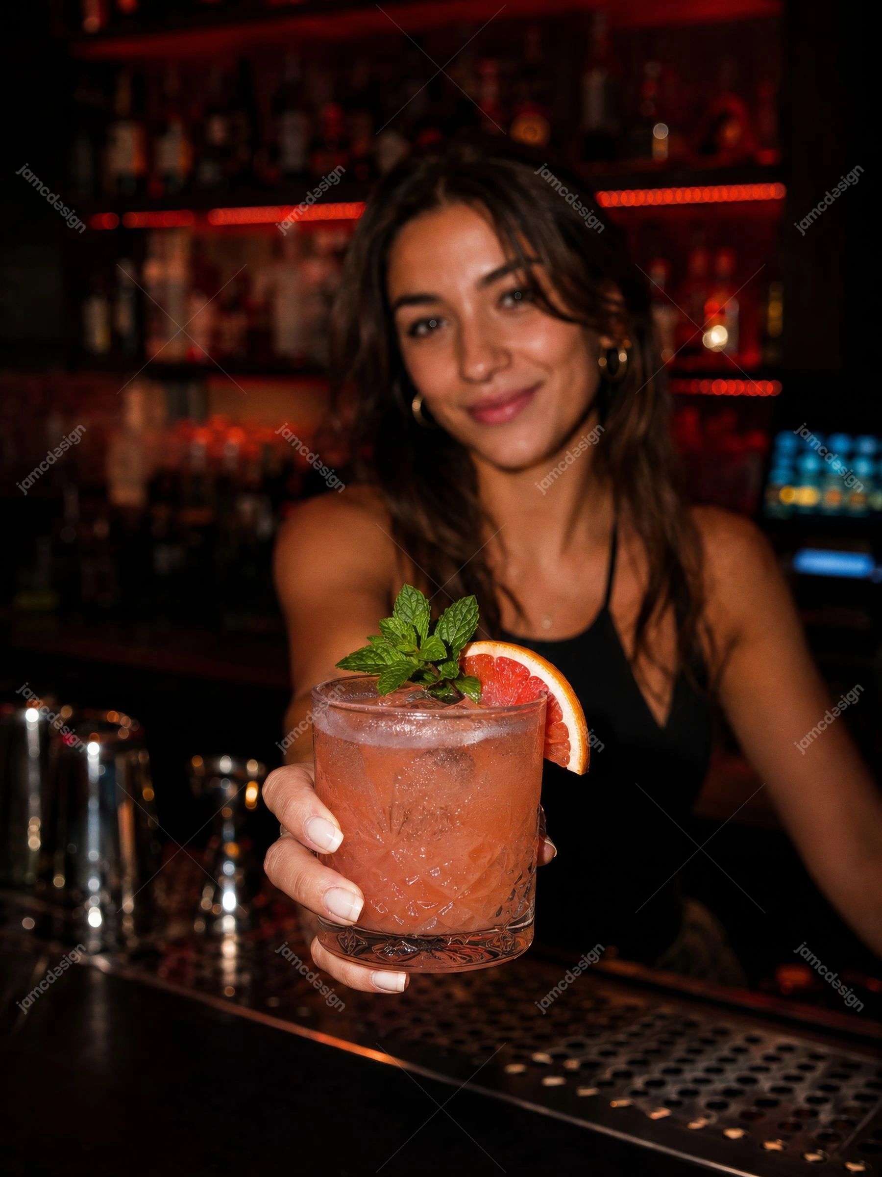 Bartender Serving Pink Grapefruit Cocktail At Night Bar. JPG photo of a woman bartender holding a pink grapefruit cocktail with mint garnish in a dim bar